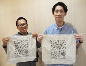 日本画家の塚原万実（依田万実）さんが日吉の街を描いたエコバッグを手に。日吉中央通り会の内田会長（左）と。右下にオリーブの木が大きく描かれている