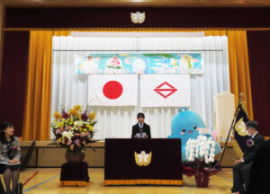 日吉台小学校キャラクター「ぶんかちゃん」をデザインした藤田さん（日吉台中学校在学中）も来場した。後ろの吊り看板は日吉台中学校の美術部が依頼を受けて制作したという