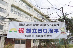 「輝け たかひが 未来へ つなげ」とのキャッチフレーズの横断幕も作られた「高田東小学校創立50周年」を祝う横断幕。キャラクターの「しばまる」が右側に描かれている（11月25日）