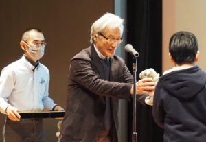 慶應義塾大学名誉教授で鶴見川流域ネットワーキング代表理事の岸由二さんが表彰状と景品の「バクちゃん」（鶴見川流域の形から生まれたキャラクター）のぬいぐるみを贈呈していた