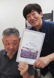 完成した「記念誌」を手に笑顔の猿渡功さん（右）。「20周年」時も編集部委員を務めた奥田徹さんが今回も編集長としての重責を果たした