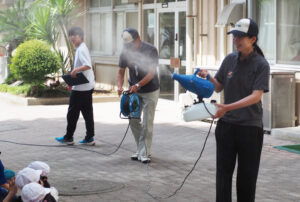 アネスト岩田株式会社（新吉田町）の渡辺さんと三矢さんが、同社の主力商品の「スプレーガン」（今回は他社商品とのこと）で暑さ対策のミストを児童に吹きかけるプレゼントも