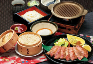 牛たん民芸定食（牛たん4枚、2390円）。「アツアツの陶板で牛たんを焼きながらお召し上がりください。焼きたての牛たんを、青唐辛子みそ・ネギ塩と一緒に食すとまた格別です。国産もち麦と国産米を使用したもち麦ご飯、北海道産とろろ、民芸自慢の手延べうどんとともにご堪能ください」と、その味わいの魅力を解説している（味の民芸のニュースリリースより）