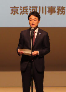 京浜河川事務所の佐々木所長が開会のあいさつ。鶴見川流域の水環境や防災に関する総合的な取り組みについて説明、下流域での河川拡幅工事や、自然環境創出のための湿地・干潟整備を進めていることを報告。綱島地区の汽水性湿地での地域学習会開催や、鶴見川流域センターでの普及啓発活動についても言及し、昨年12月に来館者数が累計50万人に達したことについても発表。来年（2027年）開催予定の「グリーンエクスポ（GREEN EXPO） 2027」に向けての環境意識醸成と合わせた取り組み拡大への意欲も示していた